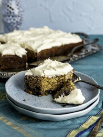 piece of cake placed in a small blue plate with a silver spoon placed on a decorative table cloth with golden stripes.