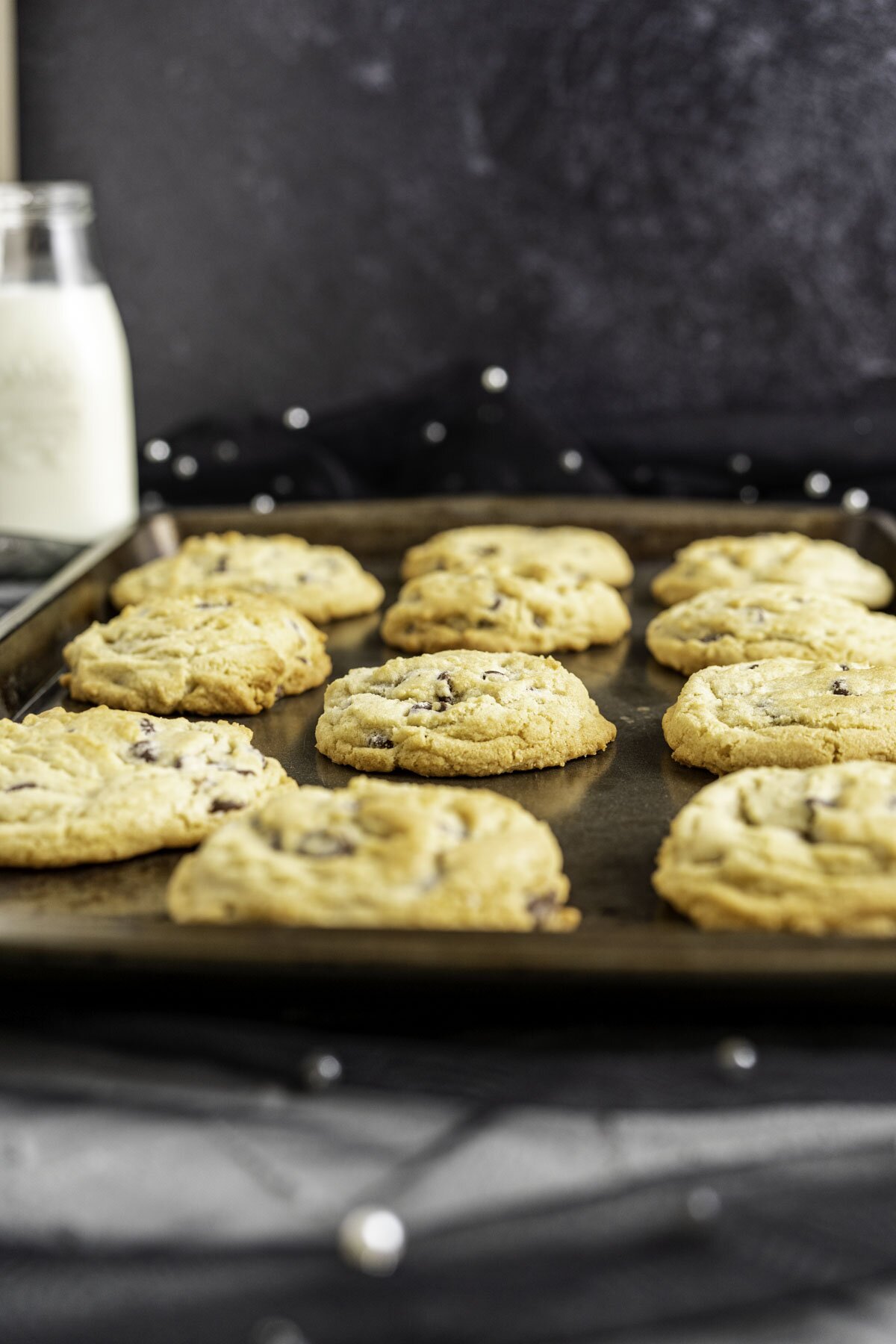 chocolate chip cookies placed in three rows in an antique cookie tray with a bottle of milk in the backdrop.