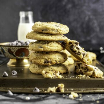 five chocolate chip cookies placed on one another with a bowl of chocolate chips and a milk bottle in the backdrop.