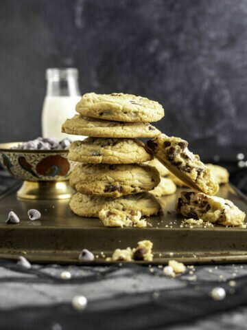 five chocolate chip cookies placed on one another with a bowl of chocolate chips and a milk bottle in the backdrop.